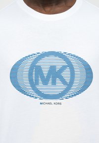 Λευκό T-shirt με μπλε οβάλ λογότυπο οριζόντιων ραβδώσεων που περιλαμβάνει διασταυρωμένα γράμματα "MK" και κείμενο "Michael Kors" κάτω από το λογότυπο.