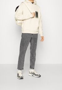 Osoba v krémovém mikině s kapsou, šedých džínech, bílých a černých teniskách, držící popruh krémového batohu přes rameno na bílém pozadí.