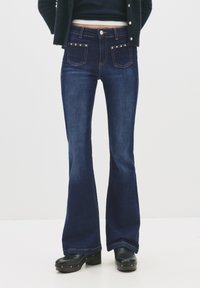 Személy magas derékú sötétkék trapéznadrágban, amelynek elején szegecses zsebek vannak, fekete magas sarkú klumpában, tengerészkék kardigánban és rövid fekete felsőben.