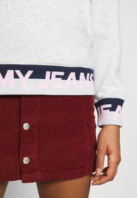 Hoodie gris con texto en navy y rosa en el dobladillo, combinado con una falda de pana roja con botones plateados. Texturas suaves y colores en contraste.