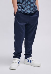 Marine sportbroek met zijzakken, gemaakt van gladde stof. Gecombineerd met witte sneakers met blauwe accenten en een geometrisch patroon shirt.