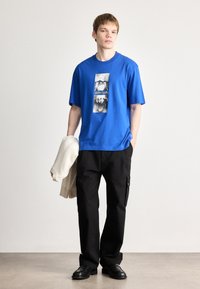 Μπλε βαμβακερή μπλούζα με γραφικό τύπωμα σε άσπρο και μαύρο. Συνδυασμένη με μαύρο cargo παντελόνι και κρατώντας μια κρέμα cardigan.