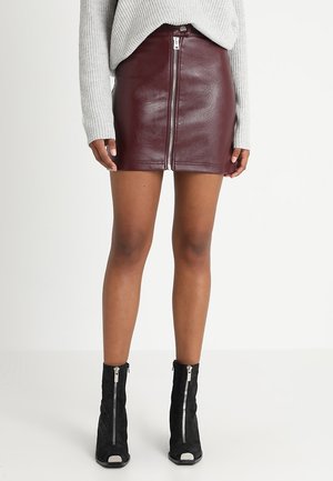 Persona che indossa una minigonna in pelle bordeaux con zip frontale, maglione grigio lavorato a maglia e stivaletti neri con zip frontale.