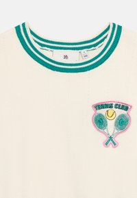 Κρεμ πουλόβερ με γιακά ριγέ σε πράσινο και λευκό, με σήμα «Tennis Club» στο στήθος που απεικονίζει δύο ρακέτες και μια μπάλα του τένις.