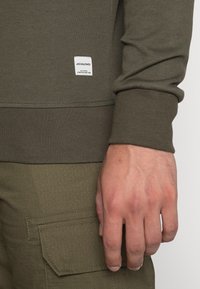 Oliwkowy sweatshirt z prążkowanymi mankietami i dołem, z białą metką marki. Dłoń opiera się na oliwkowych spodniach cargo z teksturowanym wzorem.