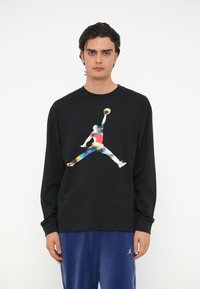 Μαύρο μπλούζα με μακριά μανίκια που διαθέτει μια πολύχρωμη γραφική απεικόνιση του λογότυπου Jumpman. Το ύφασμα φαίνεται λείο και έχει σχεδίαση με στρογγυλή λαιμόκοψη.