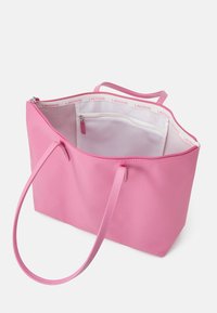 Sac fourre-tout rose en matériau synthétique lisse, ouvert en haut, avec des poignées courtes et un intérieur blanc comportant une poche zippée et un logo Lacoste.
