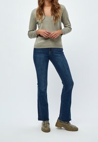 Jasnooliwkowy sweter z dekoltem w serek, zestawiony z ciemnoniebieskimi, rozszerzanymi jeansami i oliwkowozielonymi mokasynami na jednolitym tle.