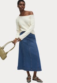 Off-shoulder krämfärgad stickad tröja matchad med en blå denim maxikjol. Outfiten kompletteras av en grön handväska och mönstrade lägenheter.