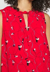 Blouse sans manches rouge avec un col en trou de serrure orné d'un nœud et de motifs floraux blancs, noirs et bleus sur un design de lignes tourbillonnantes.