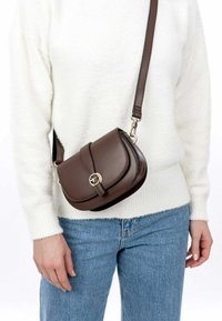 Borsa a tracolla in pelle marrone con flap curvo, accessori circolari dorati e tracolla rimovibile. Finitura texturizzata con dettagli cuciti.
