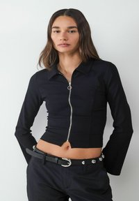 Zwarte cropped zip-up top met lange mouwen en een puntige kraag. Voorzien van zilveren rits hardware en een aansluitend ontwerp. Graagd met zwarte broek met ceintuur.