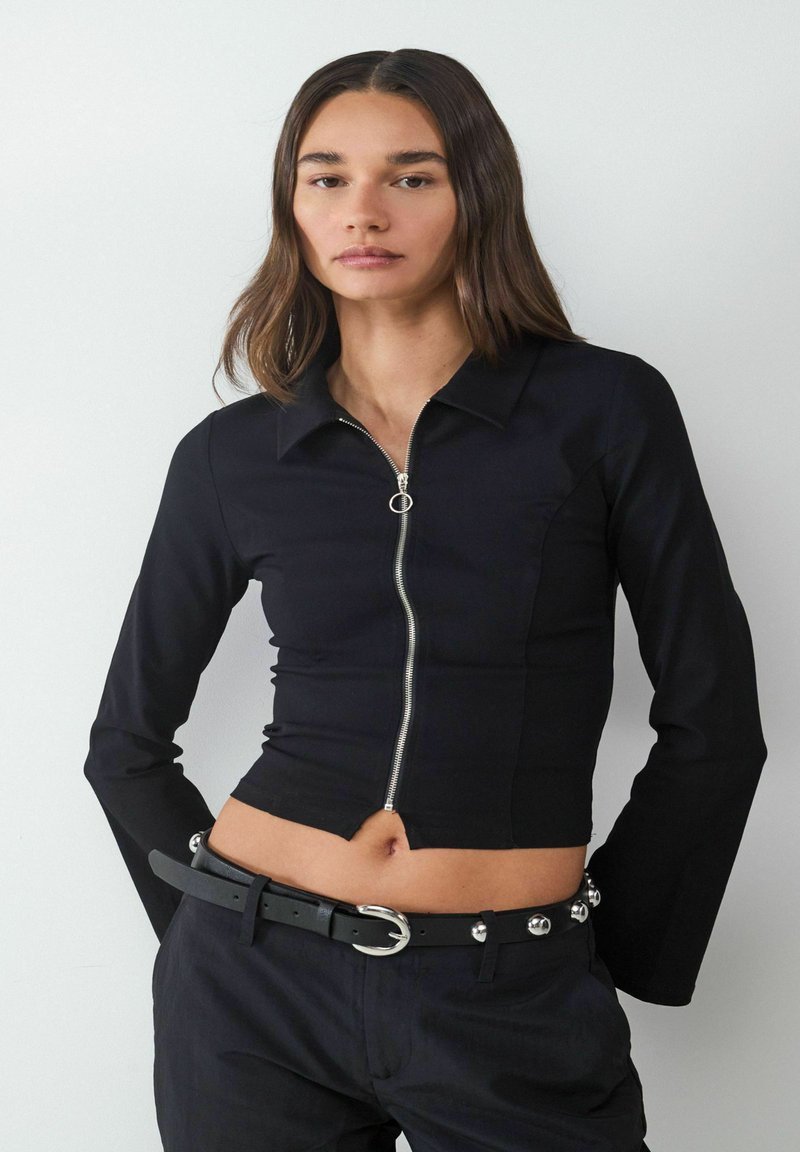 Zwarte cropped zip-up top met lange mouwen en een puntige kraag. Voorzien van zilveren rits hardware en een aansluitend ontwerp. Graagd met zwarte broek met ceintuur.