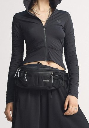 Personne portant un sweat à capuche court noir zippé, des colliers en argent superposés, un pantalon noir et un sac banane noir Adidas Skateboarding avec plusieurs fermetures éclair.