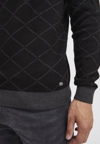 Zwart gebreid sweater met diamantpatroon, grijze accenten en geribde manchetten. Bevat een logo label aan de linkerkant nabij de zoom.