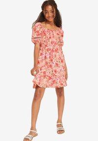 Rosa Blumenkleid mit Puffärmeln, gerafftem Oberteil, gerafftem Saum und Spitzelementen; kombiniert mit silbernen Sandalen an gebräunten Füßen.