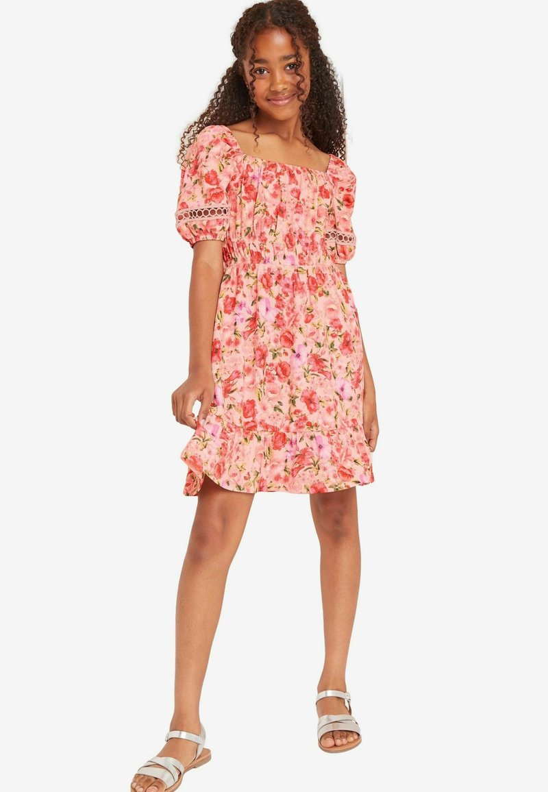 Rosa Blumenkleid mit Puffärmeln, gerafftem Oberteil, gerafftem Saum und Spitzelementen; kombiniert mit silbernen Sandalen an gebräunten Füßen.