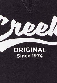 T-shirt in cotone nero con design a scollo a V. Testo bianco in grassetto "Creek" sopra "ORIGINAL Since 1974" in un carattere curvo. Texture liscia.