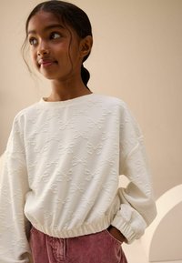 Biały, teksturowany sweatshirt z wyszywanymi wzorami w kokardy, o luźnym kroju i ściągaczach na mankietach, w zestawie z różowymi spodniami z kordurowej tkaniny.