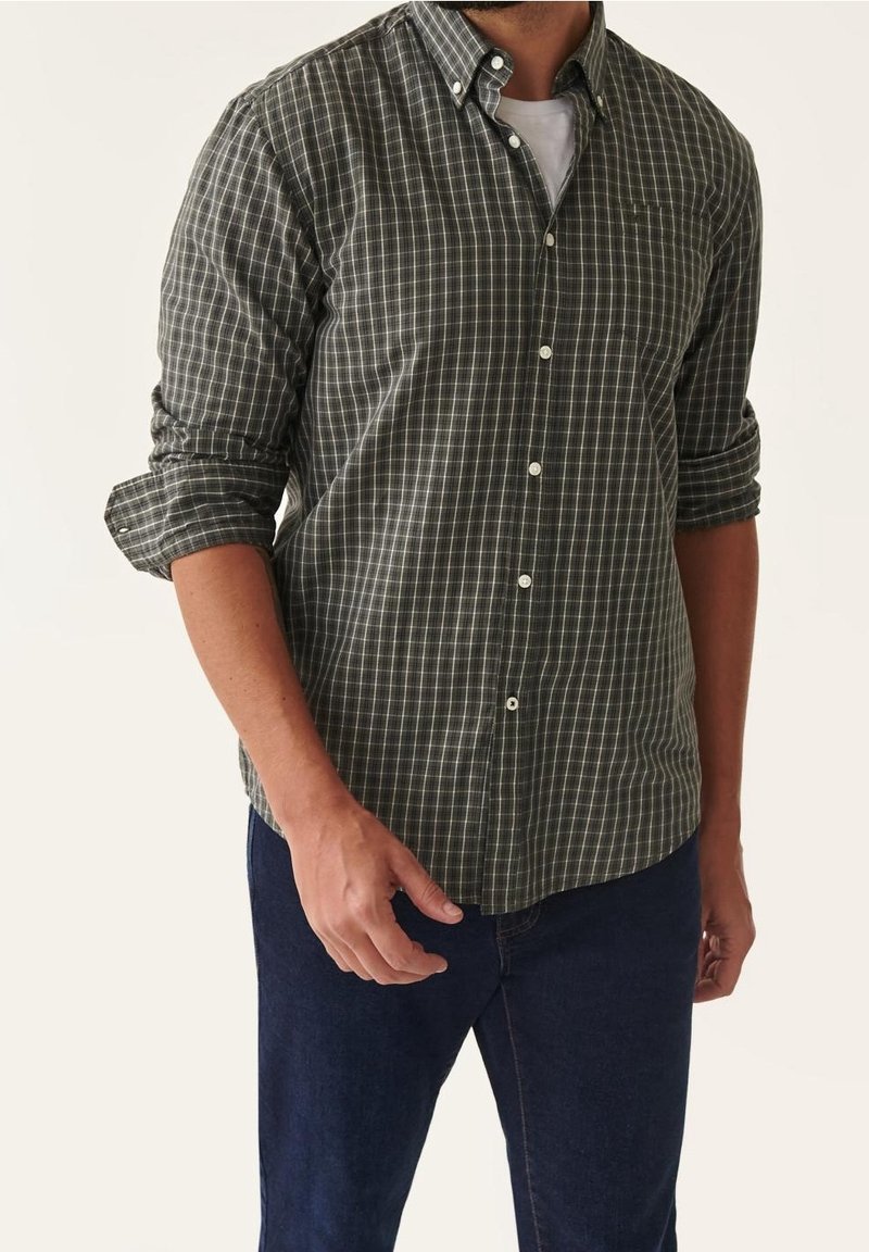 Homme portant une chemise à carreaux vert foncé et blanc à boutons avec les manches retroussées et un jean bleu foncé sur un fond uni.