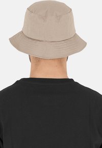 Beige spandhat lavet af bomuld, med en bred skygge og subtile syninger. Båret med en sort skjorte, set fra ryggen.