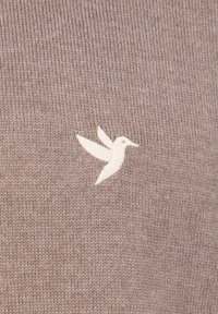 Lichtbruine gebreide stof met een wit geborduurd kolibrilogo aan de linkerkant. De textuur is glad met een fijne weving.