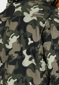 Kamouflage-mönstrad jacka i nyanser av grönt, brunt och beige. Har en slät textur, dragkedjeslutning och "SWEDEMOUNT"-logotyp på baksidan.