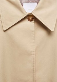 Beige trench coat featuring a large collar and a single button closure. Smooth fabric with visible stitching detail. Brand tag visible inside.