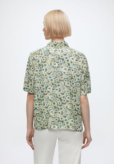 Person with short blonde hair wearing a green floral short-sleeve shirt and white pants, facing away against a plain white background.