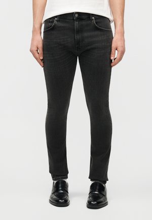 Άνδρας με στενά μαύρα τζιν, άσπρο πουκάμισο και μαύρα δερμάτινα παπούτσια, στέκεται μπροστά σε απλό φωτεινό φόντο.