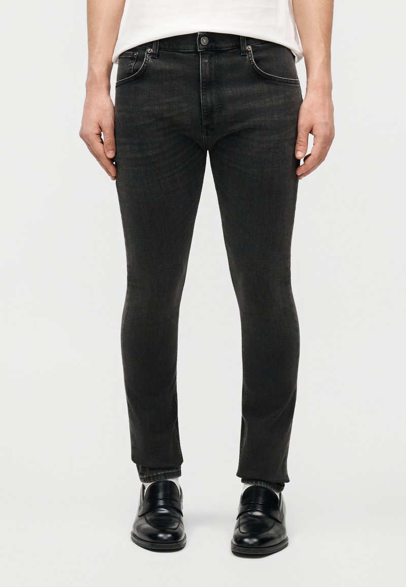 Férfi, szűk szabású fekete farmert, fehér inget és fekete bőr cipőt visel, egyszerű világos háttér előtt áll.