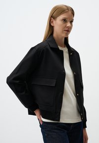 Veste tissée noire avec un col, deux poches avant, des poignets côtelés et des accents dorés. Portée par-dessus une chemise de couleur claire et un jean.