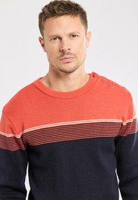 Homme aux cheveux courts portant un pull rayé rouge et bleu marine avec des boutons sur l'épaule, regardant droit devant lui devant un fond uni.