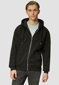 Schwarze Kapuzenjacke mit Reißverschluss aus Baumwollmischung, mit einer Fronttasche, Kordelzugkapuze und gerippten Manschetten. Trägt sie über einem hellgrauen Shirt.