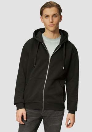 Schwarze Kapuzenjacke mit Reißverschluss aus Baumwollmischung, mit einer Fronttasche, Kordelzugkapuze und gerippten Manschetten. Trägt sie über einem hellgrauen Shirt.