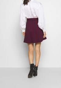 Vrouw in zwarte enkellaarsjes, een marineblauwe rok met rode verticale strepen, en een witte blouse met lange mouwen, staand tegen een effen lichte muur.