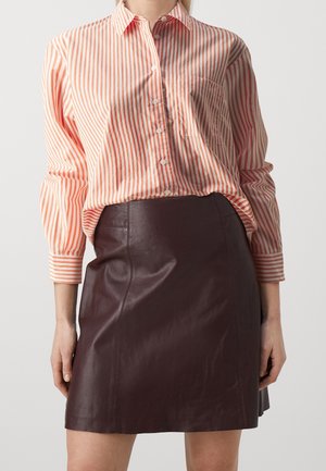 Donna che indossa una camicia a righe rosa e bianche con bottoni infilata in una gonna di pelle marrone scuro, in piedi contro uno sfondo semplice.