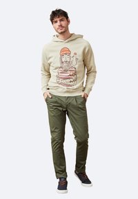 Sudadera beige con un estampado gráfico de un hombre con barba y un pájaro. Pantalones verde oliva de corte relajado. Zapatillas marino con cordones naranjas.
