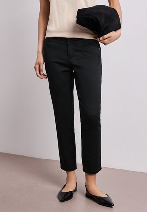 Person iført sorte slim fit bukser, beige top, sorte spidse ballerinaer, der holder en sammenfoldet sort jakke mod en ensfarvet baggrund.