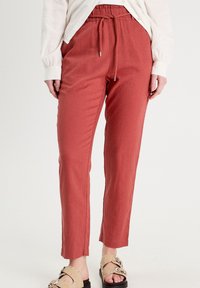 Femme portant un pantalon taille haute couleur rouille avec cordon de serrage et des sandales beiges ornées de clous, debout devant un fond blanc uni.