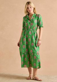 Zielona sukienka w kwiaty z dekoltem w kształcie litery V, krótkimi rękawami, elastycznym pasem, warstwową spódnicą, ozdobiona różowymi i niebieskimi wzorami, w zestawie z jasnymi sandałami.