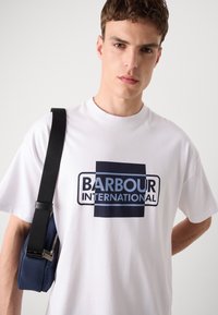 Valge puuvillane T-särk, millel on suur "BARBOUR INTERNATIONAL" logo sinise ja tumesinise värviga. Puhas lõige ümarate krae ja lühikeste varrukatega.