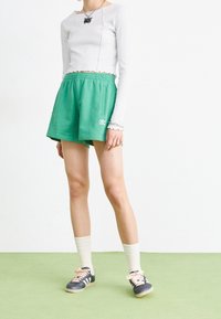 Zelené sportovní šortky s elastickým pasem, v kombinaci s bílým dlouhým rukávem. Šedé tenisky s krémovými detaily a bílými žebrovanými ponožkami.