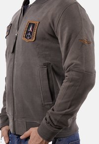 Giacca bomber grigia con zip, caratterizzata da due toppe ricamate e una tasca laterale. Il tessuto ha una consistenza liscia.