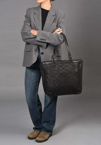 Zwarte leren tote bag met een gequiltte textuur, gedragen door een persoon in een grijze blazer en blauwe jeans, tegen een neutrale achtergrond.