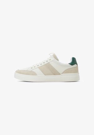 Zapatilla beige y blanca con tejido texturizado y acentos de ante, con un tirador verde en el talón, punta redonda y seis ojales para los cordones. Diseño minimalista.