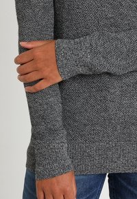 Maglione grigio lavorato a maglia con un pattern testurizzato, caratterizzato da polsini allungati. Il tessuto è morbido ed elastico, abbinato a jeans in denim blu.