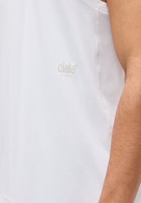 Κοντινό πλάνο στο χέρι ενός ατόμου δίπλα σε λευκή αθλητική φανέλα χωρίς μανίκια με διακριτικό λογότυπο «ciele ATHLETICS» στο στήθος.