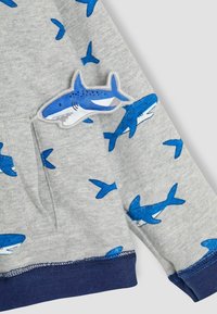 Grijze sweatshirt met blauwe haaienpatronen, met een haai patch op de zak en een marineblauwe ribbelrand. Zacht katoenen materiaal.