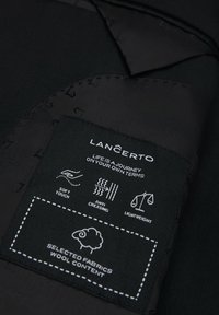 Czarny materiał z metką zawierającą białe napisy i ikony. Metka informuje o miękkim dotyku, odporności na zagniecenia, lekkiej wadze i zawartości wełny.